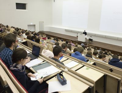 Foto: Vorlesung im vollbesetzten Audimax, am Rednerpult steht ein Professor