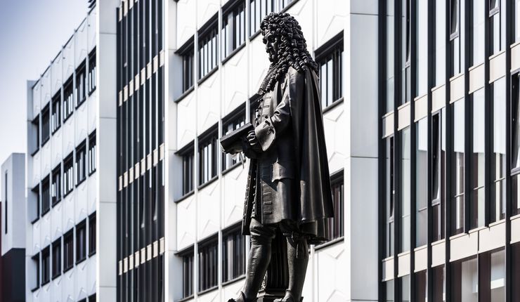Mittig im Foto ist die Bronzestatue von Gottfried Wilhelm Leibniz, die sich im Innenhof des neuen Campus der Universität Leipzig befindet. Die Statue ist nach links gewandt und komplett im Bild, ebenso wie ein kleiner Teil ihres Sockels. Hinter dem Denkmal ist die Fassade des Seminargebäudes zu sehen.