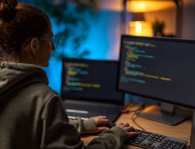 Das Foto blickt über die Schulter einer jungen Frau, die an einem Schreibtisch sitzt und auf einer Tastatur tippt. Sie trägt einen Kapuzen-Pullover und eine Brille. Auf dem Schreibtisch stehen ein Laptop und ein Desktop-Monitor. Der Text auf den Bildschirmen ist nicht lesbar, es ist aber erkennbar, dass es sich um Programmcode handelt.